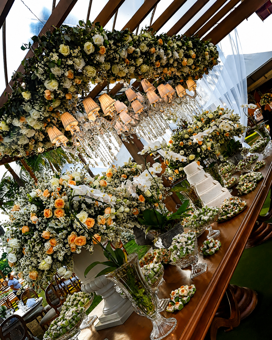 Mesa principal com decoração floral em rosas brancas e laranja sob lustres de cristal